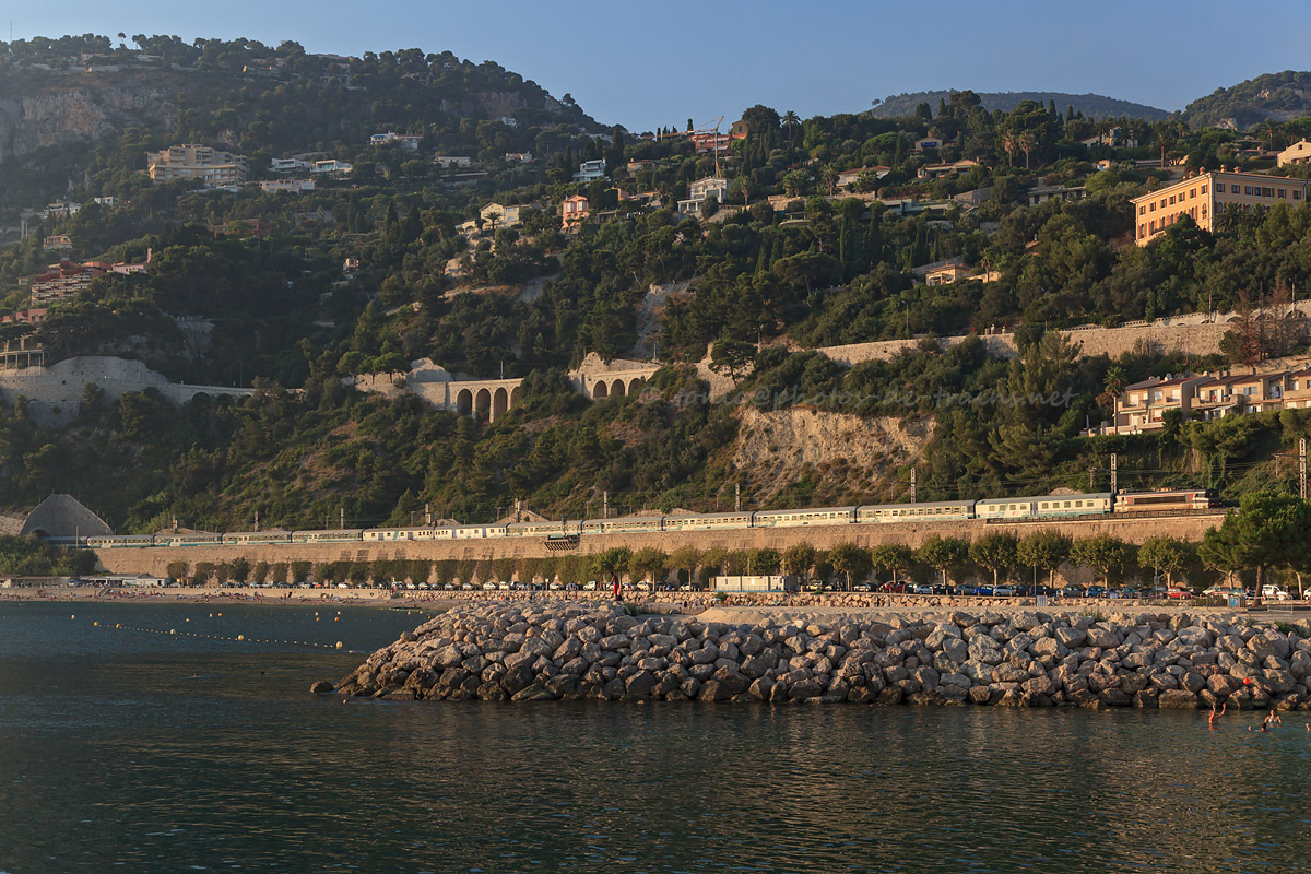 Passant dans la rade de Villefranche-sur-Mer, éclairé par un soleil déjà bas dans le ciel, le train de pèlerins 29724 Lourdes - Ventimiglia continue son trajet déjà long en direction de l'Italie. La BB 22347 est en tête.