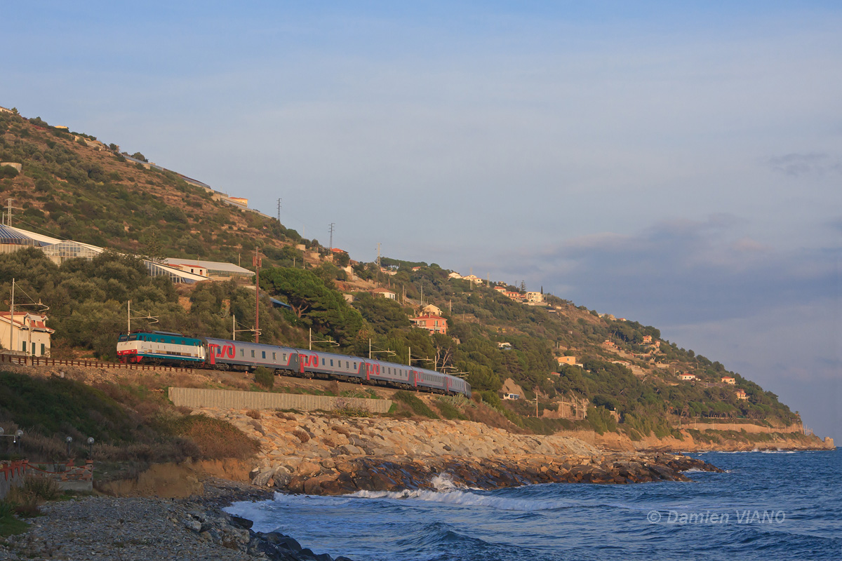Alors que l'ombre de la colline s'apprête à envahir la voie ferrée, le train 16/17 reliant la capitale Russe à la Côte d'Azur en une cinquantaine d'heures est photographié le long de la Méditerranée, après son arrêt-circulation à Imperia, sur la commune de San-Lorenzo-al-Mare. La E 444-049 assure la traction du train jusqu'à Ventimiglia, où une locomotive SNCF prendra le relais.