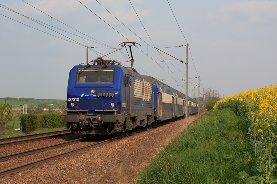 Longeant un champs de colza, la BB 27310 assure le train 165837 Paris-Montparnasse - Dreux. Elle est vue juste après la bifurcation entre les lignes de Dreux et de Mantes-la-Jolie dont on aperçoit un poteau caténaire à gauche sur la photo.